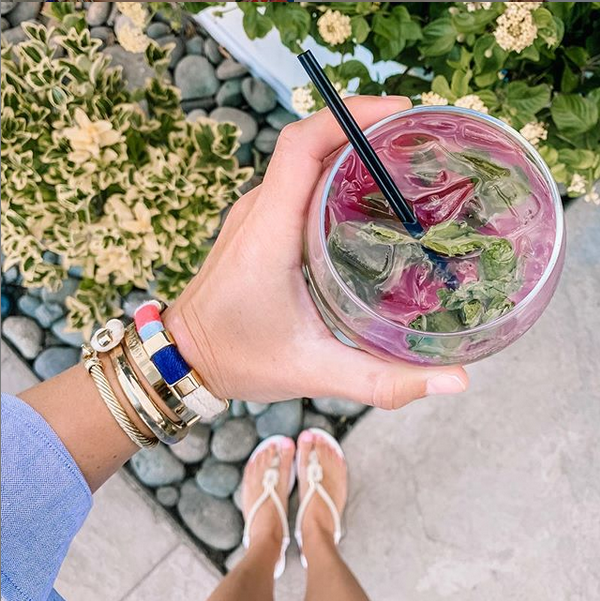 Hand holding a glass of pink iced drink with a straw, surrounded by greenery.